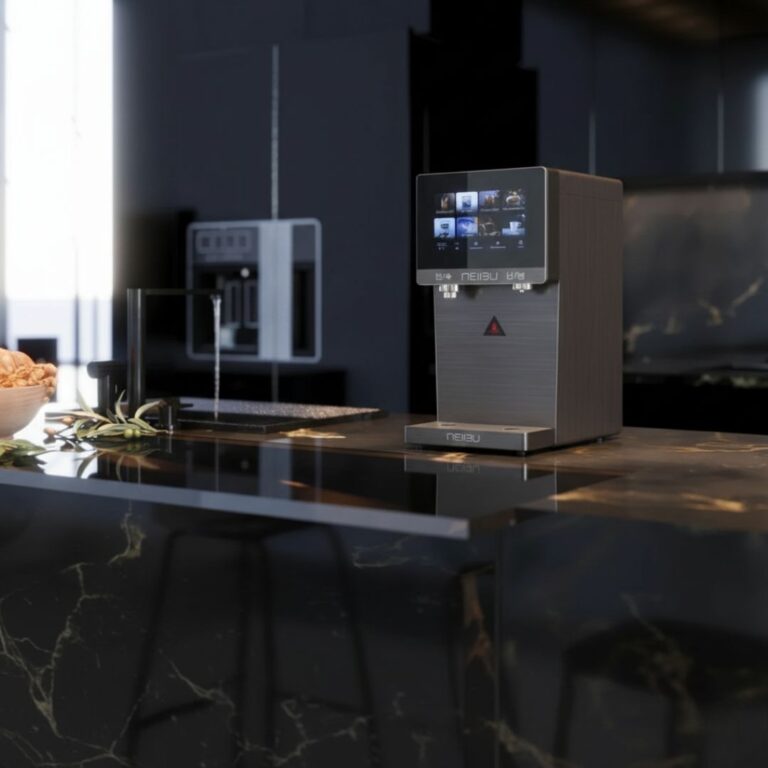 Dispensador de agua con gas de pie, de diseño vertical y color gris oscuro, marca Columbia Aqua. Cuenta con un panel de control táctil negro en la parte superior, una salida de agua central con embellecedor plateado y una bandeja de goteo ranurada en la parte inferior. El equipo está ubicado en una habitación con paredes claras, piso claro y rodapiés de madera, situado justo al lado de una estantería blanca que contiene cajitas de bambú y una cesta de mimbre en la parte superior.