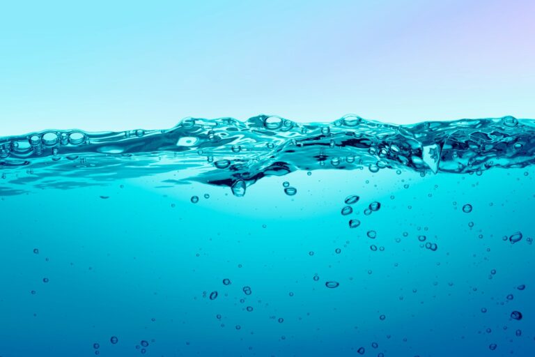 Vista transversal a nivel macro de una masa de agua en tonos azul vibrante. La línea divisoria superior muestra la superficie del líquido con suaves ondulaciones, reflejos y algunas burbujas atrapadas en la tensión superficial. En la mitad inferior, sumergidas, se observan múltiples burbujas redondas de diferentes tamaños ascendiendo por el agua cristalina, una representación visual muy gráfica para artículos que explican como se hace el agua con gas.