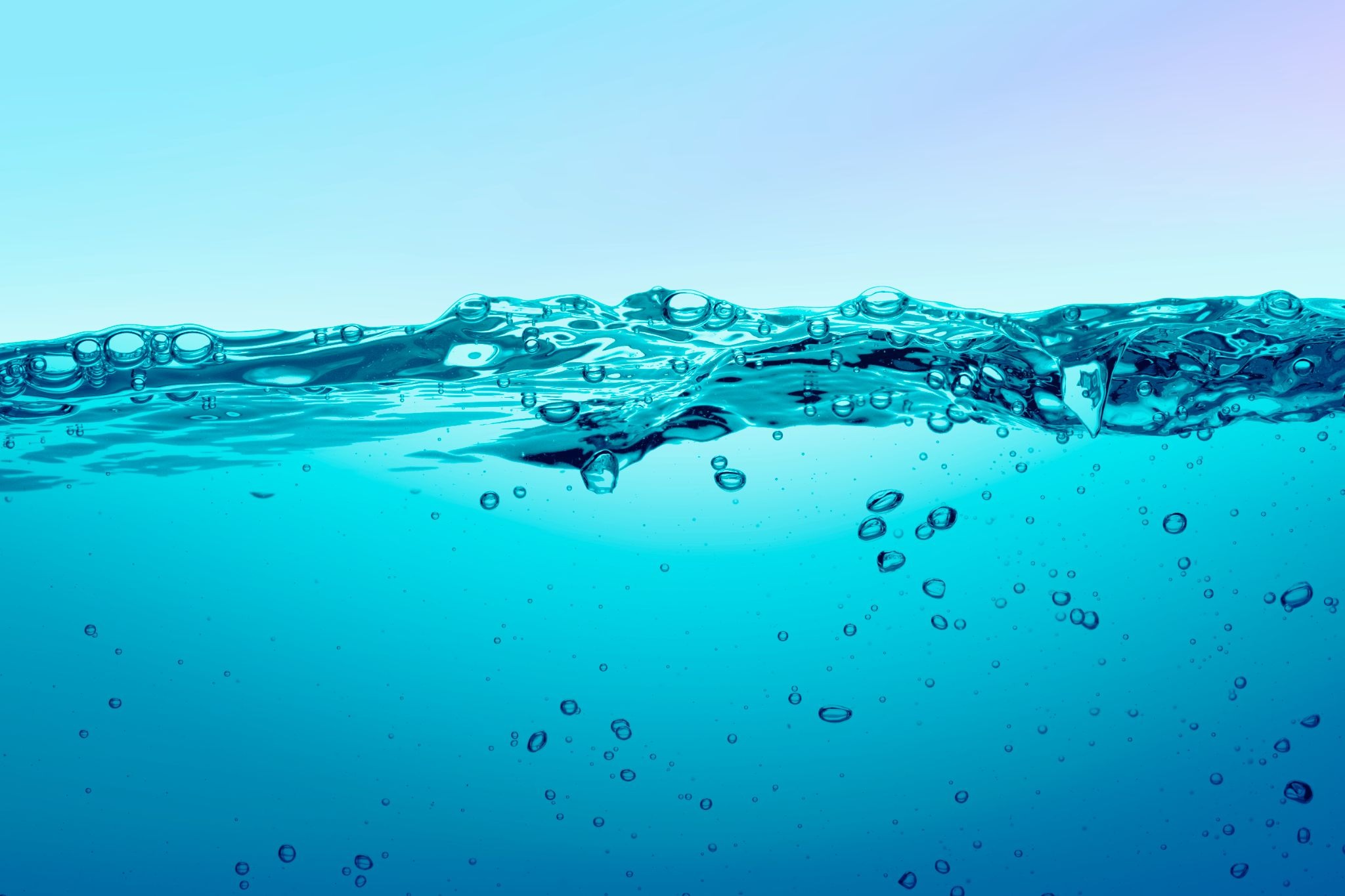 Vista transversal a nivel macro de una masa de agua en tonos azul vibrante. La línea divisoria superior muestra la superficie del líquido con suaves ondulaciones, reflejos y algunas burbujas atrapadas en la tensión superficial. En la mitad inferior, sumergidas, se observan múltiples burbujas redondas de diferentes tamaños ascendiendo por el agua cristalina, una representación visual muy gráfica para artículos que explican como se hace el agua con gas.